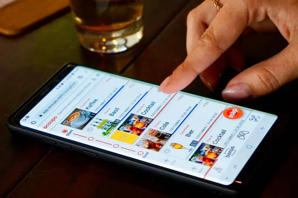 Scoupo-App in Nutzung bei einem Barbesuch Person bedient die Scoupo-App auf einem Smartphone in einer Bar, im Vordergrund ein Glas Bier und eine Hand, die auf einen Cocktail-Coupon tippt.